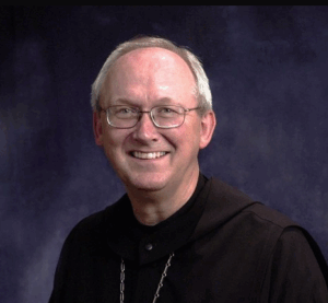 A smiling older man with glasses, short gray hair, and wearing a black robe with a chain necklace, posed in front of a dark blue background.